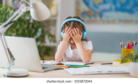 Sad Little Girl, Schoolgirl Wearing Headphones, Uses Laptop To Study At Home. Child Is Bored To Learn And Gain Knowledge Remotely. Student Wants To Go To School. Digital Concept E-learning. Remote.