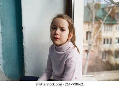 a sad little girl is sitting by the window with tears in her eyes.