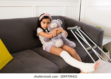 Sad little kid hugging her teddy bear and recovering from a broken leg at home. Girl using a cast and crutches