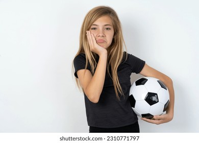 Sad lonely beautiful caucasian teen girl wearing sportswear holding a football ball over white wall touches cheek with hand bites lower lip and gazes with displeasure. Bad emotions - Powered by Shutterstock