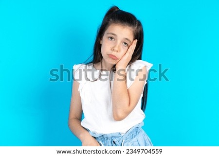 Sad lonely caucasian kid girl wearing white t-shirt touches cheek with hand bites lower lip and gazes with displeasure. Bad emotions