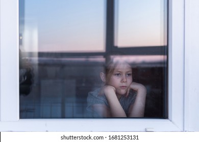 A Sad, Lonely Child Sits On The Windowsill And Looks Out The Window. Stay At Home Quarantine Coronavirus Prevention Of The Pandemic. The View From The Street. Prevention Of The Epidemic.