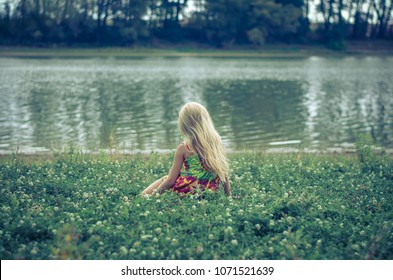 Sad Lonely Girl With Long Blond Hair In Colorful Dress Sitting Back View Alone As The Fairy In The Green Grass By The River