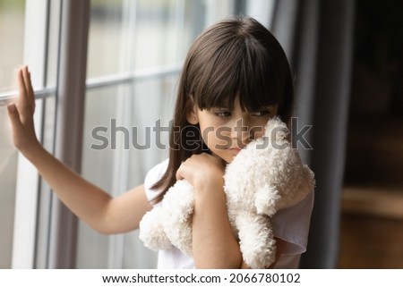 Sad lonely kid holding, hugging teddy bear toy, standing at window alone, feeling depressed, upset, unhappy, going through trauma, touching glass. Childhood problems, abuse, in family concept
