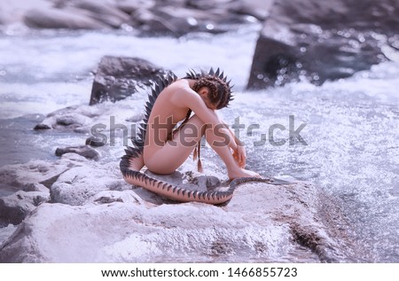 Sad lonely mermaid alien sits on the rocks by the river. Creative costume with a tail and spikes, scales all over the body. Hairstyle long hair with braids. Art photography and processing