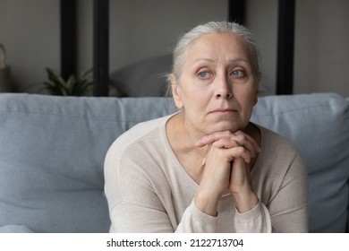 Sad lonely pensive elder lady looking away, sitting on couch at home with chin on hands, thinking over health problems, feeling sadness, boredom, apathy, concerned about memory loss, dementia
