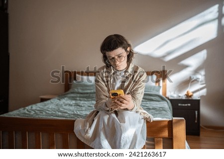 Sad lonely unhappy teenager sitting on bed, scrolling information, photos in smartphone. Reserved teen girl spending time alone at home. Addiction to social networks and internet, absence of friends