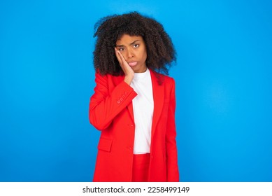 Sad lonely young businesswoman with afro hairstyle wearing red over blue background touches cheek with hand bites lower lip and gazes with displeasure. Bad emotions - Shutterstock ID 2258499149