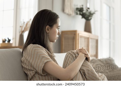 Sad lonely young woman looking away sitting on sofa at home, looks pensive, suffer from depression, personal problems, have melancholic mood, think about break up, negative emotions, stress, burnout