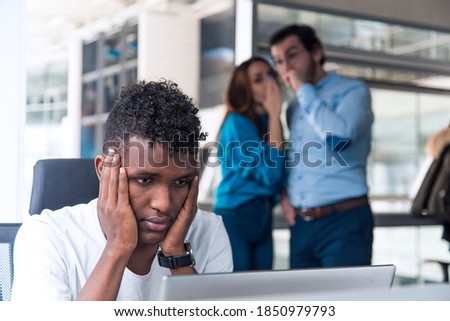 A Sad looking Black man is working on desk and there are two colleagues are gossiping at background behind of him