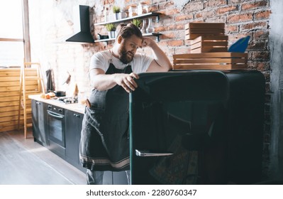 Sad male chef in white t shirt and apron staring into empty refrigerator and sighs in modern kitchen with brick wall at home - Shutterstock ID 2280760347