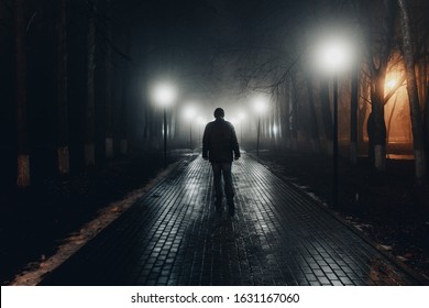 Sad Man Alone Walking Along The Alley In Night Foggy Park. Back View