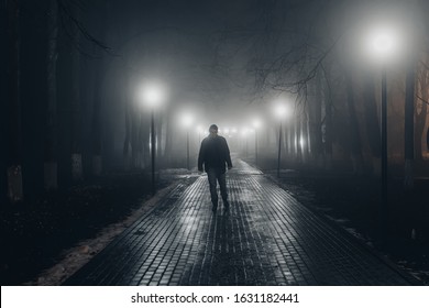 Sad Man Alone Walking Along The Alley In Night Foggy Park. Front View