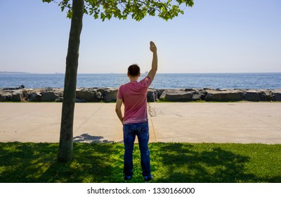 Sad Man Or Boy Standing Alone On The Seaside And Waving His Hand To Distant Ship And Horizon With Feeling Lonely. Time To Go. Missing Someone. Desire / Hope To Go Far Away. Time To Say Goodbye. 