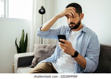 Sad man checking smartphone sitting on a sofa at home