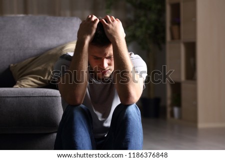 Sad man complaints sitting on the floor in the living room at home with a dark background