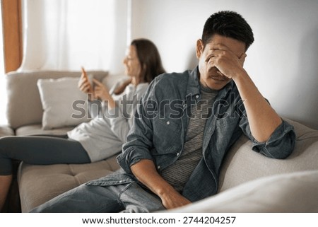 sad man covering face while partner uses smartphone on couch, relationship distance and emotional stress concept
