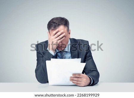 Sad man in a dark suit looks at papers and makes a facepalm, on grey background. Epic fail emotion.