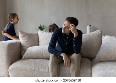 Sad Man Deep In Thoughts Looks Into Distance Sit On Sofa Fatigued Due Noisy Kids, Feels Annoyed By Running Children Play Around Him. Upbringing Problems, Divorced Single Parent In Depression Concept