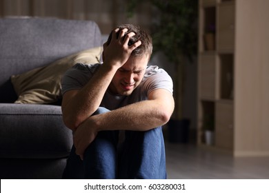 Sad man lamenting sitting on the floor in the living room in a house indoor with a dark background