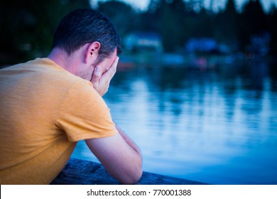 Sad Man Looking Depressed and Hopeless; Face Cupped in Hands, Crying with Back to Camera; Scenic Lake in Background (Blue Tones, Copy Space)