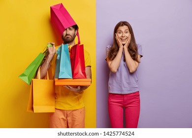 Sad man overloaded with shopping bags, has wife shopaholic, spend free time during weekend on buying new clothes, pose indoor. Positive woman rejoices successful purchases, spends money unwisely - Powered by Shutterstock