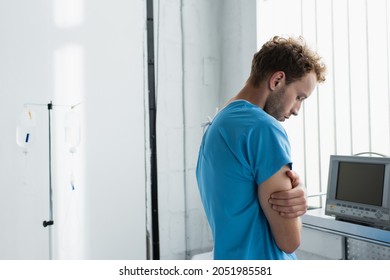 Sad Man In Patient Gown Standing With Crossed Arms In Hospital