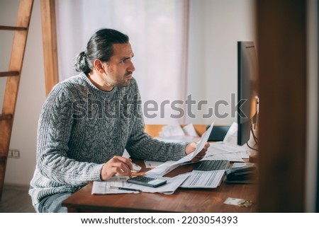 A sad man in a warm sweater is sitting at a table with utility bills in his hands.  A dejected man calculates the increased costs of heating, gas and electricity using a calculator