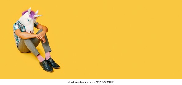 sad man, wearing a unicorn mask, sitting on the floor on a yellow background with some blank space on the right, in a panoramic format to use as web banner or header - Shutterstock ID 2115600884