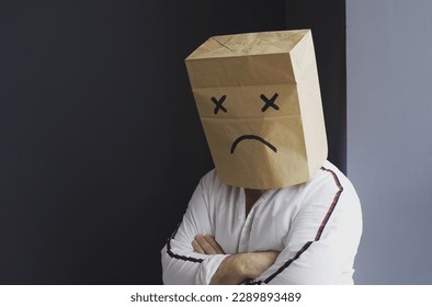 A sad man in a white shirt with a bag on his head, with a sad smiley face drawn, stands against the wall and cries. - Powered by Shutterstock