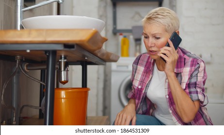 Sad mature woman calling plumber having water leaking from sink pipe. Portrait of worried aged female calling plumber crouching with basin under leaking sink pipes in bathroom
