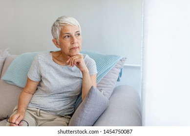 Sad Mature Woman Suffering From Agoraphobia Looking Out Of Window. Depressed senior woman sitting on sofa at home. Depressed senior woman at home - Powered by Shutterstock