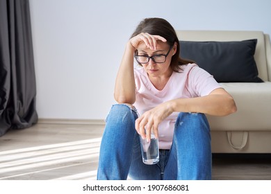 Sad middle aged woman sitting at home on the floor with glass of water. Health problems of older women, mental health, migraine pain, menopause symptom, depression period after coronavirus covid-19 - Shutterstock ID 1907686081