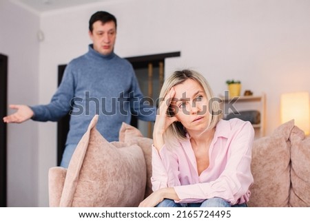 Sad middle-aged woman sits upset on the couch at home while his angry husband swears at her. Conflict in the family