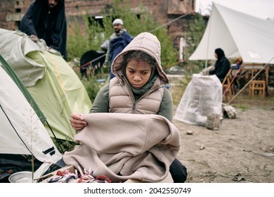 Sad Middle-eastern Refugee Girl In Hooded Vest Holding Plaid While Freezing In Tent Camp For Migrants