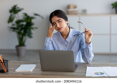 Sad Millennial Indian Female Employee Suffer From Headache, Fatigue Rubs Eyes At Workplace With Laptop In Office Or Home Interior. Stress, Deadline, Pain, Illness And Migraine Due Covid-19 Lockdown