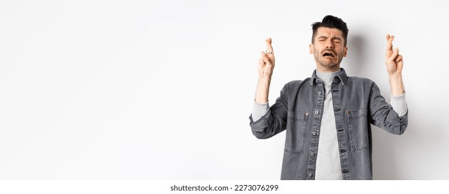 Sad and miserable man hoping god, cross fingers and crying, praying or making wish with upset hopeless face, standing on white background. - Shutterstock ID 2273076299