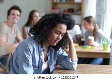 Sad Mixed Race Girl Sitting Alone Separately From Other Mates Feels Unhappy. Friends Having Fun Together In Cafe Ignoring Or Scoffing At African Peer. Outcast, Discrimination And Teen Problem Concept