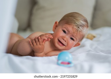 Sad Naked Almost One Year Old Blond Baby Boy Lying With His Rattle At Home On A Cozy Bed After Bathing In Prone Position And Crying Because Feeling Unwell