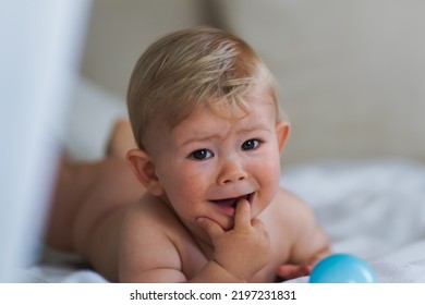 Sad Naked Almost One Year Old Blond Baby Boy Lying With His Rattle At Home On A Cozy Bed After Bathing In Prone Position And Crying Because Feeling Unwell