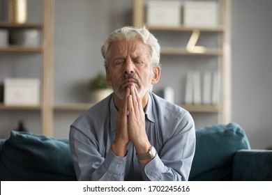 Sad Older Man With Closed Eyes Praying With Hope, Sitting On Couch At Home Alone, Holding Hands Together In Prayer, Serious Upset Mature Senior Male Meditating, Begging, Asking Help