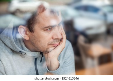 Sad Pensive Handsome Man Leaning Head On Hand In Cafe, Looking Through A Window With Reflections.