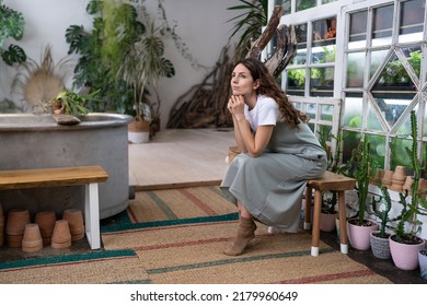 Sad pensive woman sits on bench in home garden thinking of crisis challenges looking out the window. Melancholic gardener female resting after work in floral store or greenhouse. Doubtful lady at home