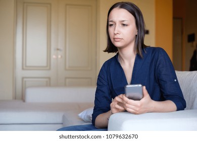 Sad pensive young girl thinking sitting on sofa holding smart phone - Shutterstock ID 1079632670