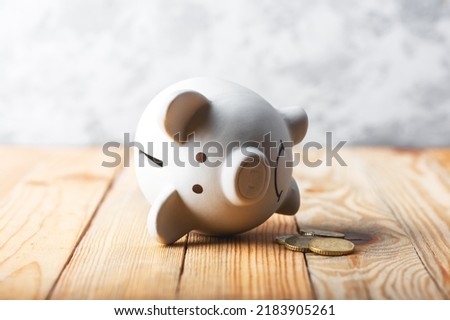 A sad piggy bank behind stack of euro coins symbolizing the fall of monetary assets. White moneybox with golden coins. Crisis concept