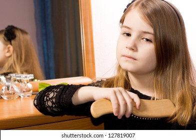 Sad Pretty Little Girl Brushing Her Long Blonde Hair With A Hairbrush Reflected In The Mirror Of The Dressing Table Behind Her