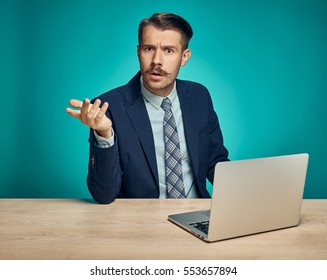 The sad and puzzled young man in a business suit working on laptop at desk on blue studio background