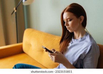 Sad red-haired young woman looking at smartphone screen expression face thinking about problem sitting on yellow couch, difficulty, feeling failure and exhausted, suffering grief and bad relationship - Powered by Shutterstock