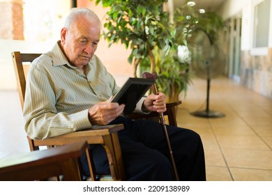 Sad Retired Old Man At The Nursing Home Looking At A Picture Frame With His Family And Missing His Partner Sitting In A Rocking Chair