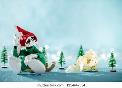 Sad Santas Helper Elf Sitting On Sledge Looking At Broken Christmas Bauble. North Pole Christmas Scene. Santas Workshop. Elf At Work. 